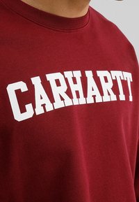 Maroon sweatshirt met vet, witte "CARHARTT" tekst over de borst. Gemaakt van zachte stof met een klassiek ronde hals ontwerp.