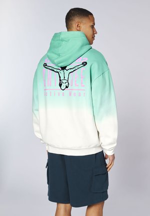 Mann trägt einen Hoodie mit Türkis-Weißem Farbverlauf und einem Taucher-Motiv sowie pinkem Schriftzug „Active Wear“ auf dem Rücken, kombiniert mit dunkelblauen Cargoshorts.