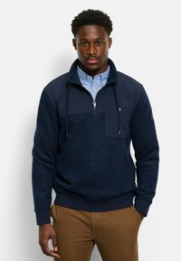 Marineblauer Pullover mit texturiertem Fleece-Körper, glattem oberen Teil, Reißverschluss am Hals und verstellbarem Kordelzug. Mit einer Fronttasche.