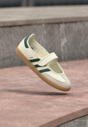 Scarpa bianca in pelle con strisce verdi, punta arrotondata, fissata da una fibbia. Suola in gomma, design minimalista, cuciture in rilievo lungo i bordi superiori.