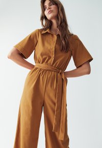 Hnědý overal s krátkými rukávy, předními knoflíky, páskem v pase a širokými nohavicemi, vyrobený z hladkého materiálu.