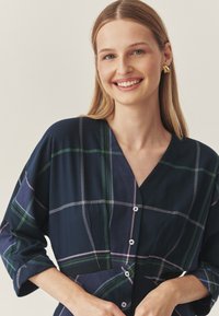 Kék kockás ruha zöld, fehér és rózsaszín csíkokkal. V-nyakú, háromnegyedes ujjak, és gombos részletek elöl. Simaságot árasztó anyagszerkezet.