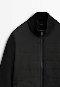 Veste matelassée noire avec col et manches côtelés, fermeture éclair à l'avant et lignes de coutures visibles sur fond blanc.