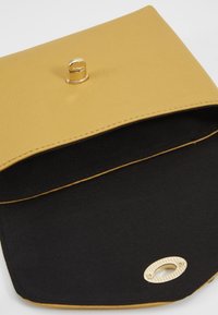 Żółta skórzana kopertówka o gładkiej fakturze, z złotym zapięciem na zatrzask i czarną podszewką wewnętrzną, wyposażona w metaliczny detal.