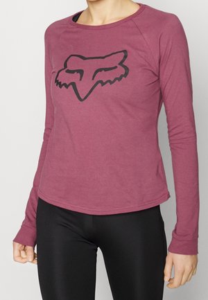 Persona con una camisa de manga larga color burdeos con un logo negro estilizado de cabeza de zorro en el pecho y pantalones negros.