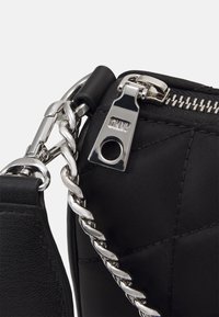 Svart quiltad väska med silverfärgad kedjeslinga, silverdragkedja med ingraverat "DKNY" och ett metallklipp som fäster selen.