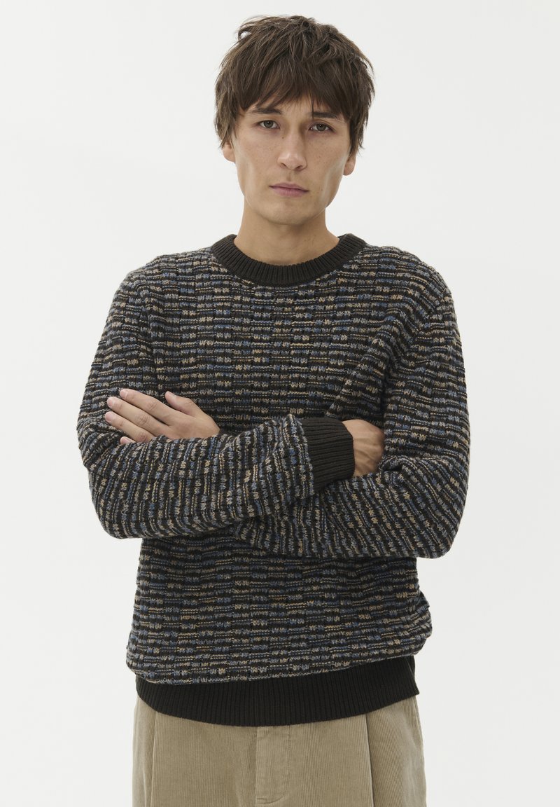 Mlad moški s kratkimi rjavimi lasmi, oblečen v pulover z rjavimi, modrimi in črnimi vzorci ter bež hlačami, stoji z rokami križanimi.