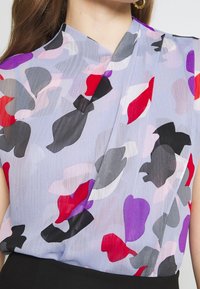Blusa ligera y transparente con una base de lavanda, que presenta un patrón abstracto en formas rojas, negras, moradas y grises. Diseño de escote en V.