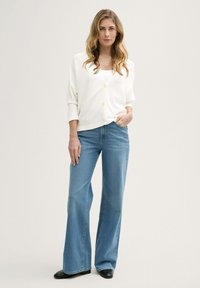 Witte knoopcardigan met driekwartmouwen en een ronde hals, gecombineerd met lichtblauwe flared jeans en zwarte enkellaarzen.