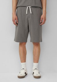 Graue Baumwollshorts mit einem Kordelzugbund, seitlichen Taschen und einem entspannten Schnitt. Getragen mit weißen Socken und beigen Sneakers.