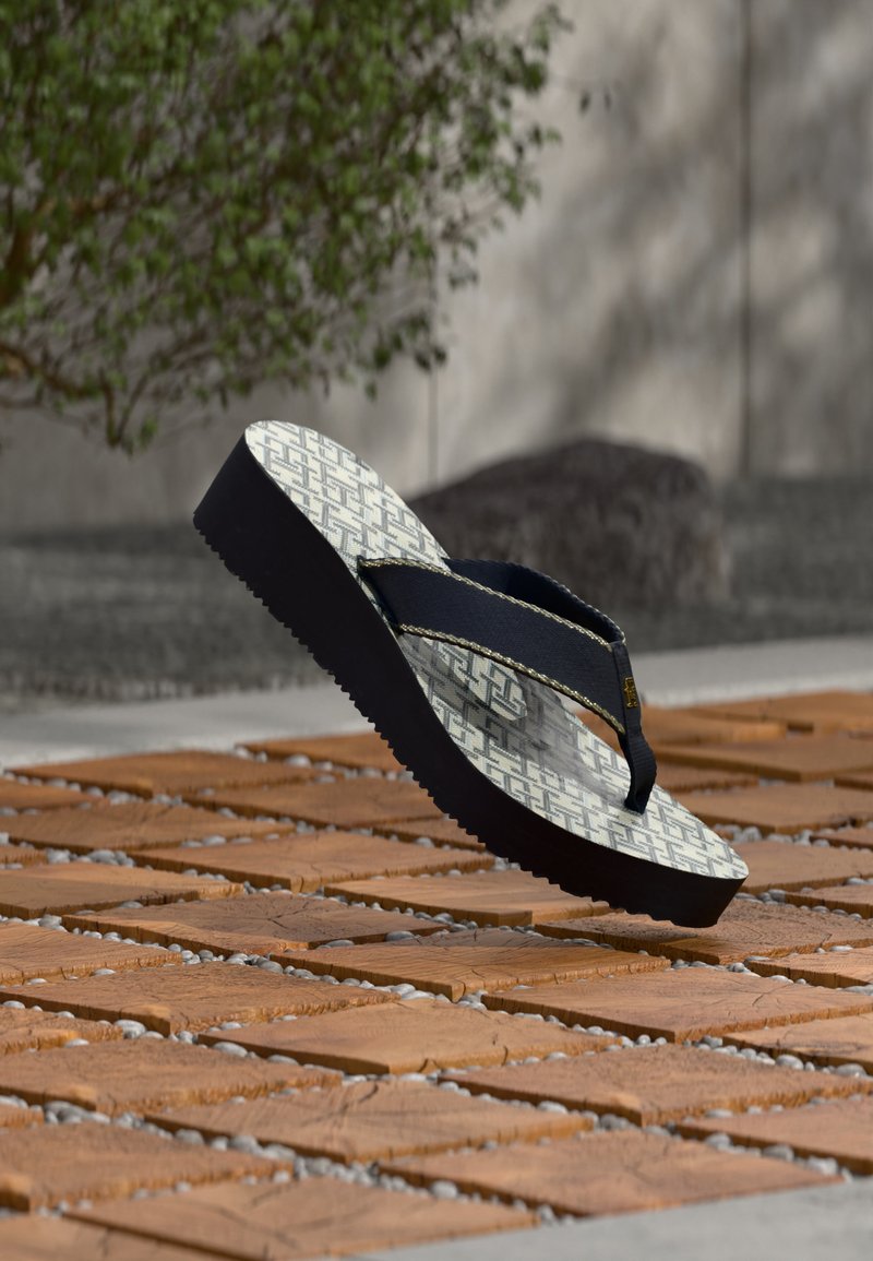 Πλατφόρμα σανδάλι flip-flop με ασπρόμαυρο μοτίβο και παχιά σόλα σε πλακόστρωτο έδαφος, με φυλλωσιές και πέτρινο τοίχο στο φόντο.