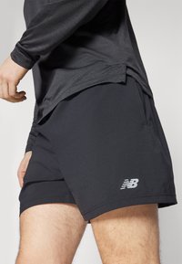 Calções atléticos pretos com uma textura suave, apresentando um bolso lateral e um logotipo refletor no canto inferior esquerdo.