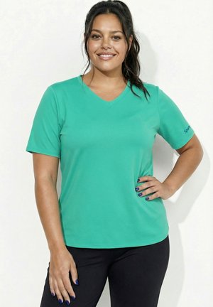 Smilende kvinde med mørkt hår i en teal V-hals T-shirt og sorte bukser står med den ene hånd på hoften mod en ensfarvet baggrund.