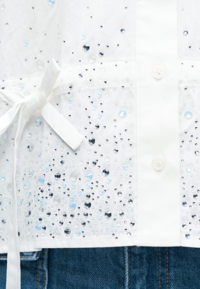 Tissu blanc parsemé de petits et grands strass clairs et argentés, patte de boutonnage, ruban blanc noué, et denim bleu en dessous.