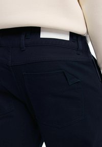 Jeans blu scuro con un design attillato, dotati di una piccola tasca a ribalta, cuciture in vista e un'etichetta bianca semplice in vita.