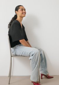 Musthave mustade mustade ja hõbedaste kingadega hall vastupidav särk, kergesti lühikesed denim ja punased kontsakingad. Modell istub hõbedasel toolil, valge seina taustal.