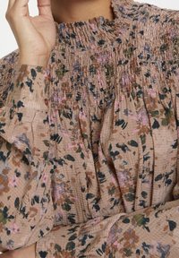 Blouse fleurie en tissu doux avec un design plissé. Caractérisée par un col froncé, des manches longues et une palette de couleurs pastel comprenant du rose, du vert et du marron.