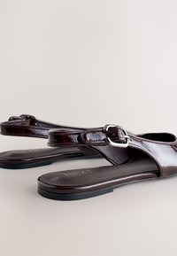 Sandales slingback en cuir verni marron foncé avec boucle argentée et semelles plates noires sur fond clair.