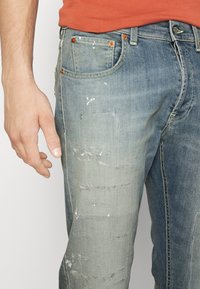 Helle blaue Denim-Jeans mit Whisker-Effekten, abgenutzten Patches und ausgewaschenen Stellen. Kupferne Nieten und gelbe Nähte akzentuieren das Design.