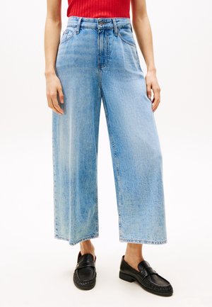 Personne portant un jean large bleu clair court, un haut côtelé rouge et des mocassins en cuir noir, debout avec les bras détendus.