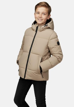 Jongen draagt een beige donsjack met zwarte ritssluiting en capuchon, handen in de zakken, glimlachend, staand tegen een effen lichte achtergrond.