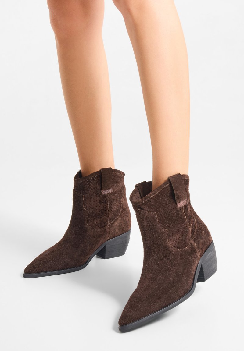 Donkerbruine enkellaarsjes van suède met een puntige neus, een gestructureerde afwerking en een korte, gestapelde hak. Voorzien van decoratieve stiksels en trekriempjes.