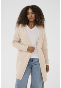 Moteris su garbanotais plaukais, vilkinti šviesiai rudą atvirą megztinį, baltą V formos iškirpimo marškinėlį, mėlynus džinsus ir sidabrinę apyrankę ant riešo.