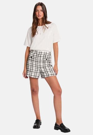 Femme debout portant un haut blanc à manches courtes et un short taille haute à carreaux noirs et blancs avec des mocassins noirs.