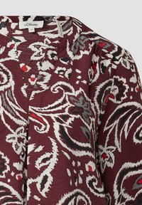 Bordeauxfarbene Bluse mit einem floralen Paisleymuster in Weiß, Rot und Schwarz. Mit Rundhalsausschnitt und strukturiertem Stoff. Markenlabel sichtbar.