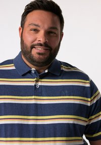 Poloko s krátkým rukávem v modré barvě se žlutými, bílými a červenými pruhy. Má límeček, tři knoflíky a mírně texturovanou tkaninu.