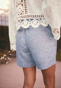 Osoba v světle modrých lněných šortkách a bílém krajkovém topu stojí venku se slunečním světlem v pozadí.