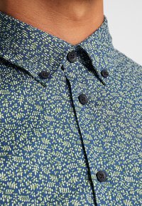 Camisa azul con un diseño de hojas en verde y blanco. Presenta un cuello abotonado y botones negros. Textura de tela de algodón.