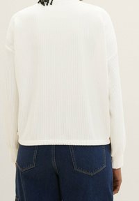 Maglione bianco texturizzato con maniche lunghe, polsini a coste e orlo arrotondato, abbinato a pantaloni in denim blu scuro con tasche posteriori.