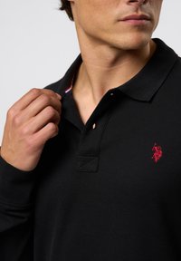 Svart polo-skjorte med teksturert stoff, med en rød brodert logo på brystet og stripete detalj inni kragen.