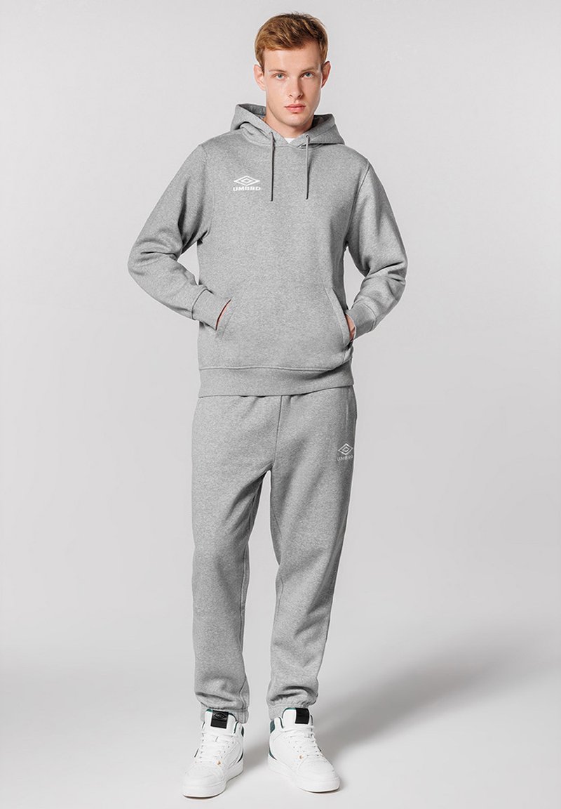 Sweatshirt gris avec capuche et poche kangourou, associé à un pantalon de survêtement gris assorti. Les deux sont dotés d'un logo, complétés par des baskets blanches.