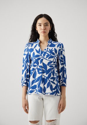 Blå og hvit mønstret bluse med V-hals, 3/4 ermer og rynkede mansjetter, laget av et lett materiale. Brukt sammen med hvite ripped jeans.