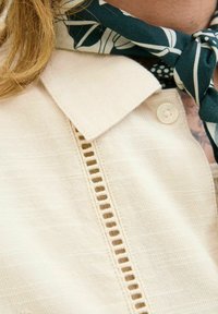 Chaqueta beige con un tejido texturizado, que cuenta con un cierre de botón, un collar y un detalle decorativo recortado en la parte delantera.