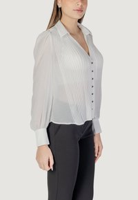 Blusa bianca plissettata con colletto, chiusura a bottoni e maniche lunghe a palloncino. Presenta una texture liscia e polsini bianchi a contrasto.