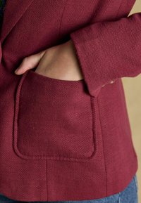 Burgundia herringbone'i mustriga ülikond, millel on sakiline krae, esine klapiga tasku ja sile tekstuur. Varrel on nupp.