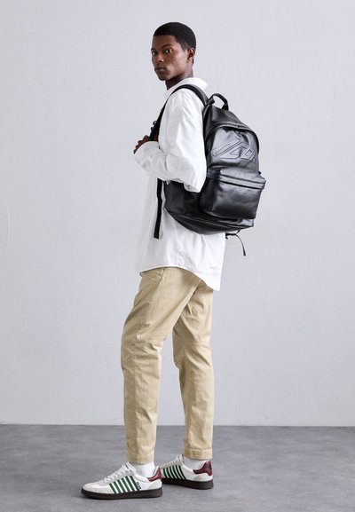 Black leather backpack with embossed logo, adjustable straps, front pocket. Model wears white shirt, beige pants, and green/white sneakers.