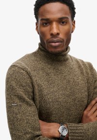Maglione a collo alto in maglia marrone con superficie strutturata, caratterizzato da un accento logo nero sulla manica. Il modello indossa un orologio rotondo.