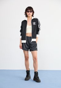 Svart Adidas-jacka med vita accenter och logotyp, krämfärgad crop top, grårandiga shorts och svarta snörstövlar. Släta texturer genomgående.
