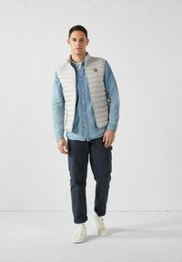 Gilet grigio imbottito sopra una camicia di denim blu, abbinato a pantaloni blu scuro e sneakers bianche. Il gilet presenta un logo e una texture liscia.