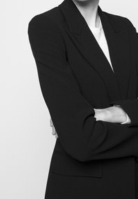 Czarna dopasowana marynarka z szerokimi klapami, wyraźnymi ramionami i gładką fakturą. Posiada jedną przednią kieszeń oraz schludną sylwetkę.