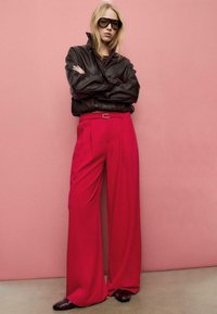 Giacca di pelle marrone, pantaloni rossi a gamba larga, scarpe scure, occhiali da sole oversize; in piedi davanti a uno sfondo rosa.