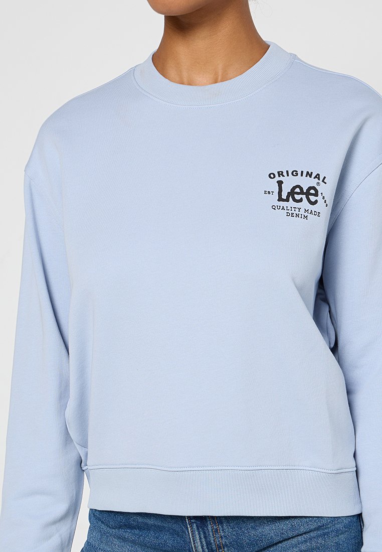 Jasnoniebieska bluza z miękkiego materiału, z okrągłym dekoltem i czarnym graficznym logo na piersi. Krótki krój oraz ściągacze przy mankietach.