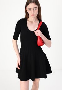 Melna, ribota mini kleita ar īsām piedurknēm, ar apaļu kakla izgriezumu un izplestu svārku. Modelis tur mazu sarkanu rokassomiņu.