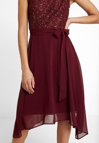Ärmlös burgunderröd klänning med spetsliv, genomskinlig asymmetrisk kjol och tygbälte knutet i en rosett vid midjan.