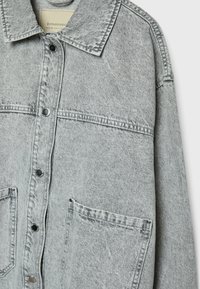 Šedá denimová bunda s límcem, předním zapínáním na knoflíky, dvěma velkými náprsními kapsami a jemnými texturálními detaily na látce.