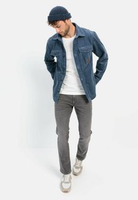 Blå fløjljakke med to brystlommer, en hvid t-shirt underneath, grå jeans og en mørk strikket beanie. Sneakers fuldender outfittet.